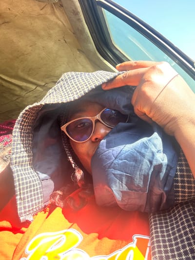 A close-up, eye-level indoor portrait depicts an individual with dark skin situated inside a vehicle. The subject wears light-framed sunglasses reflecting blue light and an orange shirt with a yellow/white graphic. A dark blue and white small-checkered hooded garment covers the head, its crumpled dark blue lining obscuring the lower face. A dark-skinned hand is visible on the upper right, holding the hood, with a thin red string on a finger. The upper left background reveals a light-colored, worn vehicle ceiling. The upper right background shows a vehicle window, through which a clear blue sky is visible. Strong direct sunlight illuminates the subject from the right, creating significant lens flare. The individual looks directly at the viewer. Visual evidence for the specified location in Rimau, Nigeria, is absent.