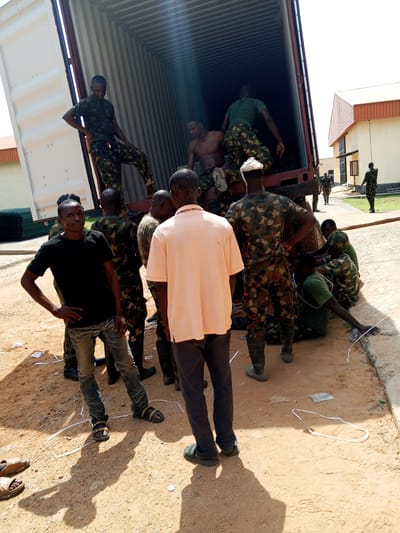 A group of individuals stands on a dirt surface in what appears to be Pasere, Nigeria. Some are dressed in military camouflage uniforms, while others are in civilian attire. A man in a black t-shirt and jeans stands in the foreground, looking toward the camera with his hand on his hip. Behind him are other individuals, some wearing camouflage, gathered around the back of a large, open shipping container that is tilted at an angle. Some of the people are positioned inside the container, while others are standing or sitting outside of it. White string-like material is scattered on the ground around the group. Buildings are visible in the background.