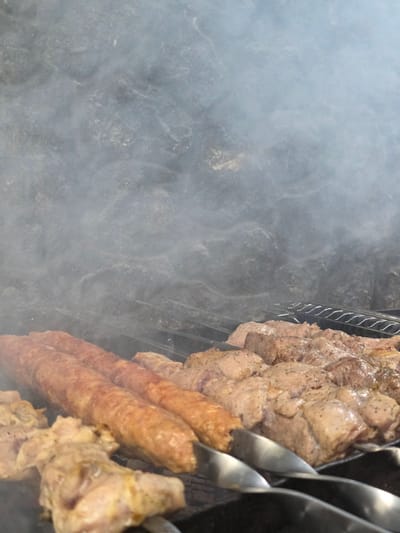 The media file shows a close-up, low-angle shot of meat cooking on skewers over a grill, partially obscured by smoke. The meat appears to be a mix of cubed pieces and cylindrical sausages, seasoned with pepper and possibly other spices. Several metal skewers are visible, holding the meat and resting on the grill grate. The background is dominated by the hazy, grey smoke, creating a diffused effect on the dark, textured surface beneath the grill. The scene is likely outdoors, given the open-air cooking and the presence of smoke. No people are visible in the frame. The lighting suggests it could be daytime, but the exact time is not discernible due to the smoke and angle. The location is identified as Pidhorodne, Ukraine. No text is visible in the image.