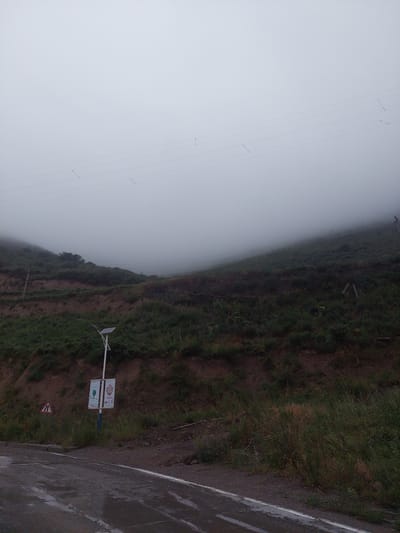 The image depicts a rural, overcast scene in Bordunskiy, Kyrgyzstan. A road, wet from recent rain, runs along the bottom of the frame, with a white line marking its edge. To the right of the road, a grassy embankment rises, dotted with dry vegetation. Further up the embankment, a solar-powered street lamp stands next to a utility pole. Two signs are affixed to the lamp post; one features a green heart symbol with text, and the other is a red sign with white text and a symbol. A triangular road sign with a red border and white interior is visible on the left side of the road. The background features a steep, vegetated hillside shrouded in thick fog, obscuring the top of the hill and the sky. Several power lines are faintly visible stretching across the foggy expanse. No people are present in the image.