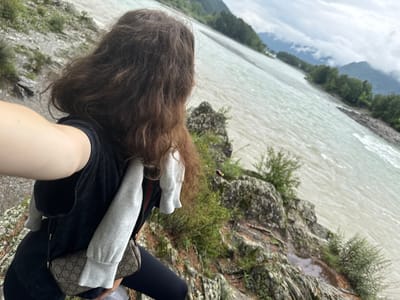 A person with long, dark, curly hair is standing on a rocky outcrop overlooking a wide, turbulent river. The person is wearing a black t-shirt with grey sleeves, dark leggings, and a small, patterned crossbody bag. The river water is a murky, light brown color and appears to be flowing quickly. The background shows a dense forest with green trees, and beyond that, mountains partially obscured by clouds. The weather appears overcast. The scene is located in Elekmonar, Russia.