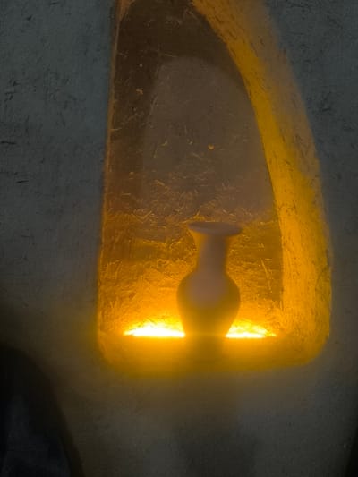 An interior scene in Turkkishlak, Uzbekistan, features a single light-colored, possibly ceramic, vase situated centrally within an arch-shaped wall niche. The vase, characterized by a flared rim and a slightly bulbous body tapering downwards, stands on a flat surface within the recess. The niche is internally illuminated from below by a bright, golden-yellow light source, casting an upward glow that highlights the rough, textured surface of the niche's interior walls. The surrounding wall, visible to the left and right of the niche, is a darker, desaturated blue-gray or dark gray, also exhibiting a rough, earthy texture. No individuals are present.