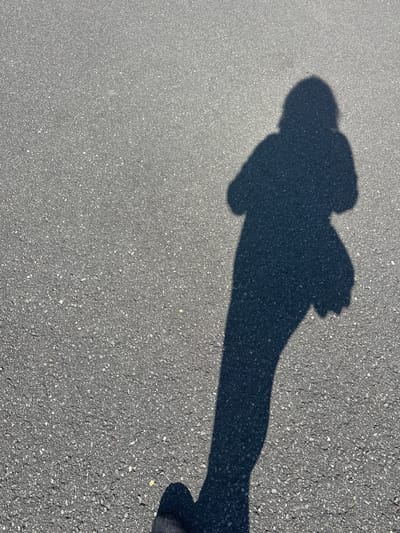 The image shows a shadow of a person on an asphalt surface. The shadow is cast from the right, with the light source high and to the right of the frame. The person appears to be wearing a hooded garment and is holding something in their left hand. The shadow's legs are visible extending down towards the bottom of the frame. The asphalt surface is dark grey and speckled with lighter aggregate. The location is identified as Ossenzijl, Netherlands, suggesting an outdoor urban or semi-urban environment. The scene is static, capturing a moment in time with no discernable action or interaction beyond the presence of the individual casting the shadow.