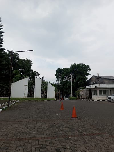 The image displays an outdoor paved entrance area under an overcast sky, located in Cianjur, Indonesia. The foreground features a dark grey and reddish-brown interlocking paver surface, upon which two orange traffic cones are positioned; one closer to the viewer and slightly right of center, the other further back and to its left. A curb with alternating black and white markings runs along the left edge of the pavement.
In the midground, a tall, dark street light pole with a horizontal arm extends from the left, supporting a small, illuminated LED light fixture. Adjacent to this pole, an entrance is defined by two white, angular architectural wall segments. Behind these walls, ornate dark metal gates are partially open, revealing lush green foliage and a white vehicle beyond. To the right of the entrance, a single-story white building, identified as a guard post or office, features windows and a grey roof with visible barbed wire. A partial sign on its roof reads "EXPRESS". A light-colored sedan is parked partially visible on the far right, near this building. Another street light pole is visible further back near the guard post. The background consists of a uniform light grey overcast sky and dense green trees. A large, dark-roofed industrial-style building is partially visible behind the guard post. No individuals are present in the scene.