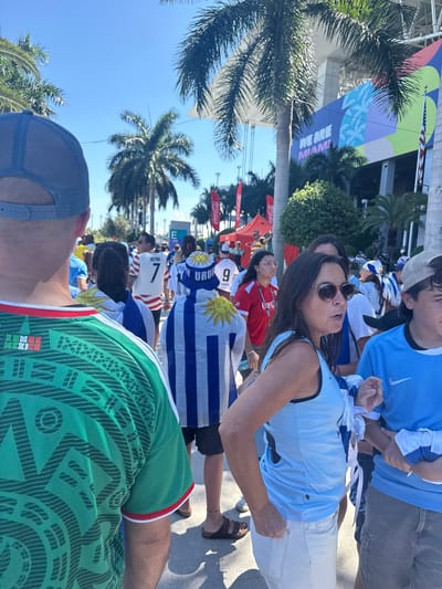 This image depicts a crowd of people gathered outdoors, likely for an event. The subjects are varied, with individuals wearing jerseys and hats, suggesting a sporting event or festival. One prominent detail is the banner in the background with text that reads "WE ARE MIAMI," indicating the location as Miami. The clear blue sky and palm trees further support a warm, sunny environment, suggesting daytime.
Several individuals are wearing what appear to be soccer jerseys with national flags or symbols. One person is wearing a light blue jersey with a Nike logo, and another is seen in a green jersey with a pattern that resembles traditional Mexican designs. Flags, including one with blue and white stripes and a sun emblem, are visible, hinting at international participation.
The overall mood appears to be one of excitement and anticipation, with people looking engaged and some appearing to be in conversation. The presence of Coca-Cola signage in the background is a common element at large public events. The image captures a vibrant atmosphere, with a focus on the fans and their attire, showcasing their support for their respective teams or countries.