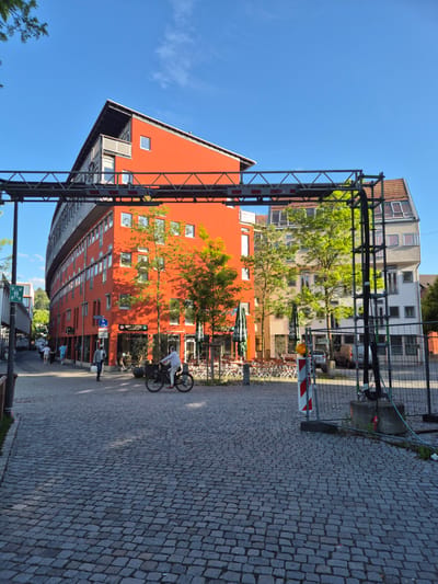 In the city of Landshut, Germany, a street scene unfolds under a clear blue sky, with the sun casting long shadows. The foreground is paved with cobblestones, leading towards a curved, vibrant orange building that dominates the left and center of the frame. This modern building features numerous windows and a distinctive slanted roofline. To the right of the orange building, a lighter-colored building with a pitched roof is visible.
In the middle ground, a person is riding a bicycle. Several other pedestrians are walking along the sidewalk in front of the orange building. Trees with bright green foliage are interspersed between the buildings, adding natural elements to the urban landscape. Outdoor seating areas with chairs and tables are set up in front of some establishments, suggesting a commercial or dining area.
A prominent metal structure, resembling scaffolding or a temporary overhead support system, crosses the image from left to right, partially obscuring the buildings and sky. A construction barrier with red and white striped markings and a concrete base is visible on the right side of the scene, indicating ongoing work. The overall impression is a dynamic urban environment with a mix of modern architecture and street life.