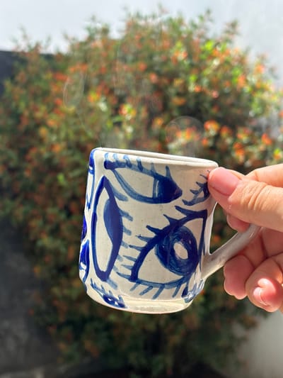 A hand holds a ceramic mug with blue stylized floral designs against a blurred background of green foliage and hints of orange or red berries. The location is likely San Juan Cuautlancingo, Mexico, although this cannot be confirmed from the image alone. The mug appears to be handmade or artistically decorated, suggesting possible local craftsmanship. The photograph is taken outdoors, with the out-of-focus background implying a garden or natural setting.