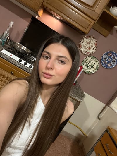 The image displays a close-up view of a young adult female subject within a kitchen, identified by the prompt as being located in Lucca, Italy. The woman has long, straight, dark brown hair parted in the middle and fair skin, looking directly towards the viewer. She is wearing a white top.

The background shows a kitchen environment. To the subject's left, a speckled grey countertop holds a four-burner gas stove with a single metal pan resting on it. A red indicator light is illuminated on the stove. Adjacent to the stove, a clear bottle and a smaller blue object are visible on the counter. Wooden upper cabinets are situated above the counter, beneath which a dark, rectangular appliance is mounted. A section of a brown wicker-patterned cabinet is visible in the lower left.

To the subject's right, a wall painted a muted reddish-brown color features three decorative wall plates arranged vertically. The top plate is white with a floral pattern, the middle is cream-colored with a green pattern, and the bottom is blue with a multi-colored floral design. An open wooden upper cabinet containing a white ceramic pot is visible to the far right. A yellow flexible hose runs along the wall at the floor junction, and a segment of terracotta-colored tiled flooring is visible in the lower right corner, along with part of a light brown wooden cabinet or drawer unit.
