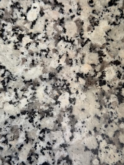 The image shows a close-up view of a speckled granite surface. The granite has a light-colored base with black, gray, and white flecks and larger crystalline inclusions. The texture appears rough and natural, typical of quarried stone. There are no people, objects, or actions depicted, and no contextual information is provided to place this granite in Arcahueja, Spain.