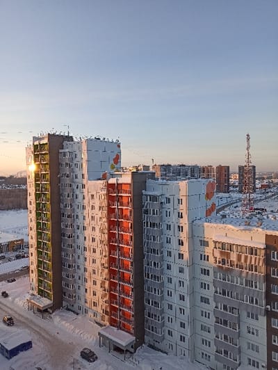 Multiple high-rise apartment buildings dominate a snowy landscape under a light blue sky. The buildings display a mix of colors, including green, red, and gray. In the distance, a construction crane and other buildings are visible on the horizon. Parking lots and roads are covered in snow surrounding the buildings. The image appears to be taken from an elevated viewpoint, offering a comprehensive view of the residential area.