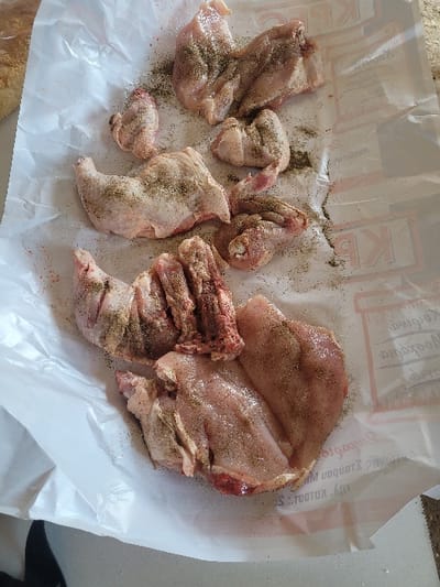 The scene captures a close-up, overhead view of raw, butchered chicken pieces seasoned with black pepper and salt, arranged on a sheet of white butcher paper. The paper prominently displays red and grey Greek text, including "ΚΡΕΟΠΩΛΕΙΟ" and "ΑΝΔΡΑΒΙΔΑ", identifying the location as Ανδραβίδα, Greece. Visible chicken components include bone-in and boneless portions, such as a thigh, breast cuts, and wings, exhibiting pink flesh and some skin. Dark specks of ground pepper and fine white granules of salt are distributed across the chicken surfaces and the surrounding paper. A blurred, light-brown object, possibly bread, is partially visible in the upper-left corner. The butcher paper rests on a light-colored, reflective surface, likely a kitchen countertop. No individuals or active interactions are present in the frame.