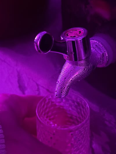 In Shebanovo, Russia, a close-up, low-angle shot captures a chrome tap dispensing a pink liquid into a textured glass. The scene is bathed in a deep purple light, likely from ambient lighting or a filter. Condensation covers the tap, indicating it is dispensing a cold liquid. The tap itself has visible markings, including the words "Open" and "Close," suggesting it is a valve for a dispenser. A hand is visible beneath the glass, holding it in place. The background is dark and indistinct, with some fabric-like texture visible in the lower left. The overall impression is one of a quiet, personal moment, perhaps of refreshment or preparation of a beverage, illuminated by striking purple hues.