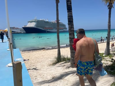 An overhead view of a beach in the City of Freeport, Bahamas shows a man in blue and yellow patterned swim trunks walking away from the camera on a sandy beach. He has a light green bag slung over his shoulder and is carrying folded clothes. In the background, a large cruise ship is docked in the turquoise water, with several other cruise ships visible further out. Numerous people are swimming and sunbathing on the beach and in the water. Palm trees are scattered across the sandy shore. The sky is clear and blue.