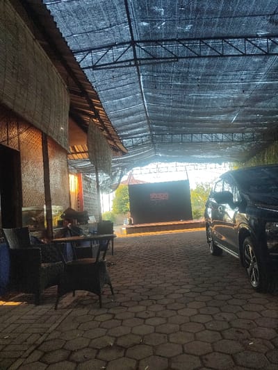 The scene takes place under a large black tarp stretched over a wooden structure. The tarp is supported by metal beams, and the structure has a thatched roof. It is likely located in a restaurant or cafe. There are four wicker chairs in the foreground and a small table in front of the chairs. A dark-colored car is parked on the right side of the scene. The paved ground is made of hexagonal concrete blocks. In the distance, there is a large black sign with the word "SOEGI" in red letters. The sky is visible through the tarp. The time of day is likely afternoon, as the sun is shining and there are shadows on the ground. The scene is in Kecamatan Bangil, Indonesia, at the coordinates -7.61 latitude and 112.772 longitude.