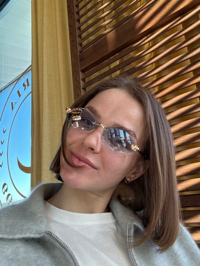 The scene, located in Mysiadło, Poland, captures a female individual from the chest up, posing for a photograph. She has shoulder-length light brown hair and fair skin, with a subtle smile and slightly parted lips. She wears rimless sunglasses with gold-colored decorative elements on the frames and temples, reflecting an interior space. Her attire consists of a light-colored top beneath a grey, textured jacket featuring a visible zipper on the left side. A small, circular earring is present in her right ear. The background shows an indoor setting: to the left, a window displays a blue sky and reflected, reversed text partially legible as "RIYA," accompanied by decorative motifs. A yellow curtain is visible beside the window. To the right, a wooden slatted structure casts distinct linear shadows onto a yellow surface, indicating direct sunlight.