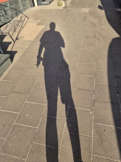 A shadow of a person stands on a paved surface. The shadow is long, indicating the sun is low in the sky. The paving stones are rectangular and laid in a pattern. There is a railing and steps to the left of the shadow. The area appears to be an outdoor public space, possibly a street or pathway. The image does not provide any specific information about the location within the city of Sanem, Luxembourg.
