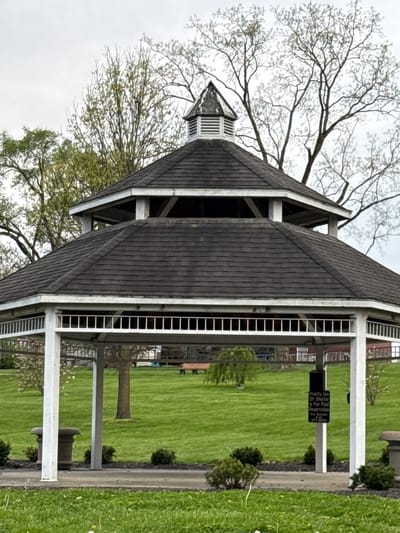 The image depicts a gazebo located in Grove City, Ohio, United States of America. The gazebo has a multi-tiered roof with dark gray shingles. White posts support the roof structure. The gazebo is surrounded by a lawn with green grass. A sign is visible that says "Priority Use Of Shelter Is For Paid Reservation. For Details 277-3000". Some shrubs are planted next to the gazebo. Trees with bare branches are visible next to the structure. A cloudy sky is overhead.