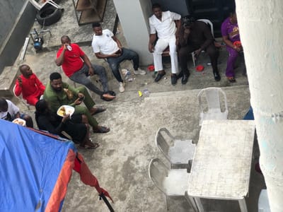 The scene depicts an outdoor gathering of nine individuals within a concrete courtyard or patio area in Aiyetoto-Asogun, Nigeria. The area is partially enclosed by a light-colored building wall.

Seven people are seated in the mid-ground and background. In the mid-ground, a man in a red shirt drinks from a clear plastic bottle. To his left, another man in a red outfit is seated. Below them, a man in an olive green traditional garment is looking at a mobile phone, while a woman in a black dress eats from a white plate containing rice and a protein. A partially visible person to the far left also holds a plate of food.

Towards the rear and right, three individuals are seated near the building wall: a man in a white traditional top and trousers, a man in dark brown attire with his hand supporting his head, and a woman in a purple patterned dress holding a small red bowl.

In the foreground, an empty white rectangular plastic table is positioned with two unoccupied white plastic chairs. The concrete ground exhibits scattered refuse, including plastic bottles and wrappers. To the lower-left, a large blue tarp partially covers an object, secured by an orange fabric tie. In the upper-left background, a discarded tire, a blue pump, and metal shelves are visible against a wall. The gathering appears to be a casual social event with food and beverages.