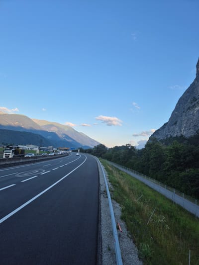 The image depicts a highway curving through a mountainous landscape under a clear blue sky with scattered clouds. On the left, a multi-lane highway features white lane markings and a speed limit sign with a circle and a diagonal line. A white truck with a crane attachment is visible on the far left, along with several cars and trucks traveling on the highway. In the distance, mountains cast shadows, and some buildings and infrastructure are present. On the right side of the highway, a steep, rocky mountain face rises prominently, covered in some vegetation at its base. Between the highway and the mountain, a walking or cycling path is bordered by a fence and flanked by a grassy area with some wildflowers and wooden stakes. The overall scene suggests a natural environment adjacent to a major transportation route. The location is identified as Kematen in Tirol, Austria.