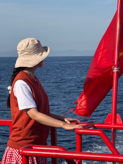 A person wearing a light-colored bucket hat, a white t-shirt, and an orange sweater vest is leaning on a red railing on the deck of a boat. The person's left hand rests on the railing, and their right hand is near their chin. A red flag with a yellow star in the center, attached to a red pole, is prominently displayed on the right side of the image, partially obscuring the view of the sea and distant hazy mountains. The sea is blue with visible waves, and the sky is clear. The image appears to be taken in Dong Son, Vietnam, suggesting a coastal or riverine setting.