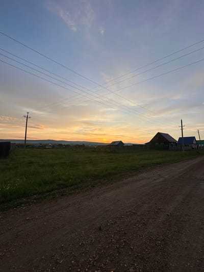 This image depicts a rural scene at dusk in the village of Urazovo, Russia. A dirt road occupies the foreground, leading into a grassy field. Several wooden houses with pitched roofs are visible in the middle ground, some with fenced yards. Power lines stretch across the sky, supported by wooden utility poles. The sky is a gradient of blue and orange, indicating either a sunset or sunrise, with scattered clouds. In the distance, rolling hills or mountains are faintly visible against the horizon. The overall impression is one of peaceful, rural tranquility. There are no people or animals visible in the image.