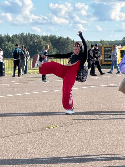 The image depicts a group of people engaged in some form of exercise or demonstration outdoors. The primary subject is a young woman in the foreground, dressed in a black top, red track pants with white stripes, and white sneakers. She is in a dynamic pose, with one leg extended and her arms outstretched, seemingly mid-movement. Her facial expression suggests concentration or exertion.

Behind her, several other individuals are visible, mostly in athletic or casual wear. Some appear to be in similar poses, while others are standing or watching. They are positioned on a paved surface with visible white lines, likely a track or an open area for activities. In the background, there is a line of trees and a blue sky with scattered white clouds, indicating daytime. A yellow structure with "ENERGY GROUP" written on it is partially visible in the upper left portion of the image.

The setting appears to be an outdoor recreational area, possibly a sports facility or a public space designed for group activities. The lighting suggests it is a sunny day, likely midday. The overall atmosphere seems to be one of active participation and engagement in a shared event. The crops highlight individual figures and the textures of the ground and sky.