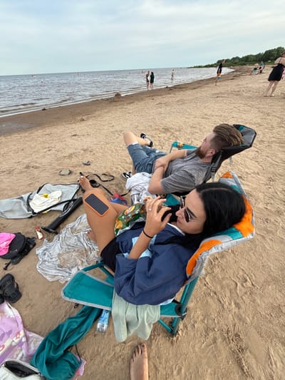 The image captures a couple relaxing on a sandy beach in Ondvor, Russia. The scene is set by the gentle lapping of water on the shore and the vast expanse of the sky. The couple is seated in beach chairs, appearing to be taking a break from their day. The woman is looking down, possibly at the bag of chips she is holding, while the man is looking away, his arms crossed. Scattered around them are various beach essentials, including a water bottle with visible text "ISOL," a phone with an orange case, towels, and bags. The presence of other beachgoers in the distance suggests a leisurely, possibly summer day. The lighting indicates it could be daytime, and the overall atmosphere seems relaxed and casual. The details in the image suggest a typical beach outing, with no specific event being the central focus.