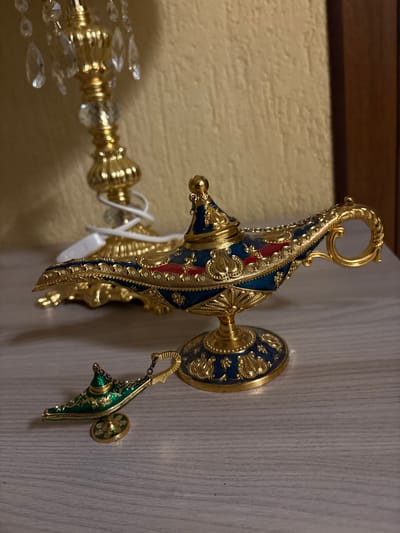 The image displays an indoor still life in Mikrevo, Bulgaria. A prominent gold-colored decorative lamp, styled as Aladdin's lamp with intricate dark blue and red enamel details, stands on a light-toned wooden surface. This lamp features a pointed lid, a curved spout, an ornate handle, and rests on a matching circular base. To its lower left, a smaller, green and gold-colored Aladdin's lamp charm, attached to a delicate chain, also rests on the same surface. In the blurred background, a tall, gold-plated decorative lamp with multiple clear faceted crystal pendants is visible, with a white power cord wrapped around its base, positioned against a textured light beige wall. A dark brown vertical furniture element is present on the far right background. No individuals or dynamic actions are depicted.