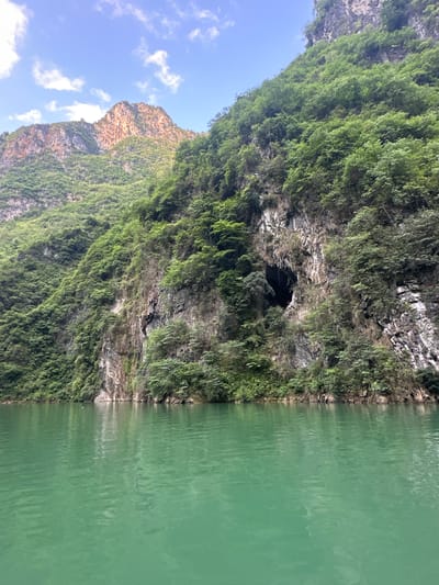 An eye-level, tilted outdoor shot shows a mountain covered in lush green vegetation bordering a body of water. In the middle of the shot, a dark, cavernous opening in the rock is partly concealed by greenery. The body of water, which occupies the right third of the image, has a green tint and appears calm, showing some reflection of the mountain. The mountain covers most of the image, with its jagged peak visible on the left side, against a blue sky with sparse white clouds. The photo gives the impression of a hidden, natural location in the Đồng Văn Commune of Vietnam.