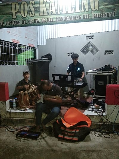 This image captures a musical performance or practice session by three men in what appears to be an outdoor or semi-outdoor setting, possibly a community security post (Pos Kamling) in Baleendah, Indonesia, during the evening or night.

In the foreground, a man with a receding hairline is seated, playing a white electric guitar with concentration. To his left, another man, wearing a cap, sits on a concrete step, skillfully playing two traditional Indonesian drums, resembling kendang. In the background, a younger man stands, focused on playing a keyboard synthesizer. He wears a dark polo shirt with reflective details on the sleeves.

The scene is set with various musical instruments and sound equipment, including two large speakers (one visible on the left, another partially on the right), amplifiers, and numerous cables running across the concrete ground. An orange and black bag lies open on the ground near the guitarist. The artificial lighting suggests the event is taking place after sunset.

Above the performers, a banner is prominently displayed, which reads: "POS KAMLINGU KP. CIPICUNG RT. 02 RW. 03 KEL. MANGGAHANG KEC. BALEENDAH." This text provides precise location details within Baleendah, Indonesia. To the far left, a window secured with a metal grille is visible, with the word "DR.SONNE" printed twice on its frame. The musicians appear engaged and focused on their performance, creating a dedicated yet casual atmosphere.