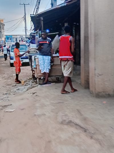Three men are standing on a dusty street corner. The first, closest to the foreground, wears a bright orange shirt. The second, standing beside a white vehicle, has on a patterned shirt. The third man, farthest away, wears a red top and light-colored shorts. There are buildings and electrical wires visible in the background. The scene appears to be a street scene in a developing area.