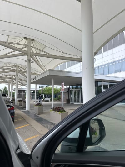 The image depicts the entrance to the "Maternity & Infant" section of a medical facility, likely a hospital, in Hoffman Estates, United States of America. The scene is captured from the perspective of someone inside a vehicle, with the car's interior and side mirror prominently in the foreground.
The building's exterior features a modern architectural style with a large, white tensile fabric canopy extending over the entrance, supported by white cylindrical pillars. Behind the canopy, a multi-story building with a facade of reflective windows and horizontal louvers is visible. The entrance itself has a covered walkway leading to revolving glass doors.
Outside the entrance, several elements are present:
* A "No Parking" sign is positioned on a pole.
* A person in a dark uniform, possibly a healthcare worker or security guard, is standing near the entrance, wearing a face mask and holding what appears to be a clipboard or tablet.
* White benches are placed along the walkway.
* Large concrete planters contain dark red and purple flowering plants.
* A black car is parked to the left of the image, with yellow lines indicating parking spaces on the asphalt ground.
* A metal grate runs along the edge of the walkway.
The overall environment suggests a functional, well-maintained public space. The lighting appears to be overcast, indicating a daytime scene. The presence of the car interior suggests the image was taken by a person arriving at or departing from the facility.