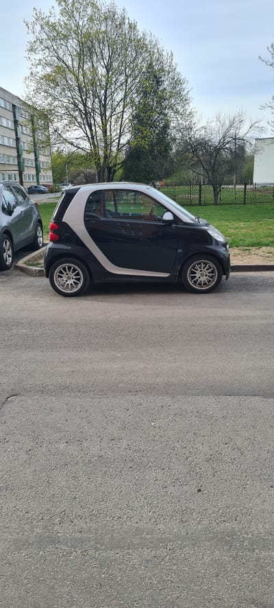 The image shows a black and white Smart car parked on the side of a paved road in Kohtla-Järve linn, Estonia. The road is concrete and shows signs of wear. A green grassy area is located beyond the car, with trees and a building visible in the background. A second car is partially visible near the Smart car. The sky is overcast.