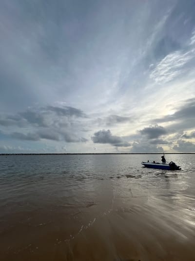 A wide shot captures a serene water scene, likely a harbor or bay in Colombo, Sri Lanka. The foreground is dominated by a wet sand shore, where gentle waves create ripples and foam along the waterline. In the midground, the calm water stretches towards a distant breakwater constructed of large stones, forming a straight, horizontal line. A small, blue and white motorboat with an outboard motor is positioned in the water, slightly right of center. Two figures are visible in the boat; one is standing, appearing to steer or operate the vessel, while another is seated. The sky above is a mix of dramatic clouds and patches of bright sunlight, suggesting either sunrise or sunset. The overall atmosphere is tranquil and the lighting is soft, indicating overcast or twilight conditions.