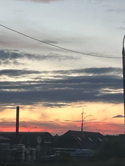 The image depicts a dusk sky with layered clouds of varying shades of gray and white, transitioning to warm hues of orange and red near the horizon. A power line runs diagonally across the upper portion of the frame. Below the sky, silhouetted structures include buildings with dark roofs, some featuring skylights, and a tall, thin pole or chimney on the left. Streetlights with curved arms are visible above the buildings. In the foreground, a stack of what appear to be construction materials or large packages is partially visible, suggesting an industrial or commercial area. The setting appears to be an outdoor scene at twilight, possibly on the outskirts of a town or city. The location is identified as Novoguslevo, Russia.