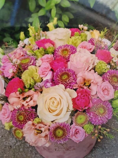 This is a close-up, top-down, studio shot of a flower arrangement in a pink box. The arrangement contains roses, dahlias, carnations, and other assorted flowers in shades of pink, cream, yellow, and green. The flowers are tightly packed, creating a lush and vibrant display. The background is blurred and appears to be a mix of green foliage and a dark surface, possibly a table or countertop. The box is made of a soft, matte material and has an ornate, embossed design. The overall impression is one of a beautiful and carefully curated floral gift. There are no people visible in the image. The image does not provide any information about the city of Vigneux-sur-Seine, France.