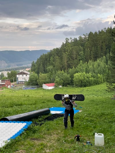 A person lies on the ground, on their back, with a snowboard attached to their feet. They are wearing a helmet, goggles, and dark-colored snowboarding attire. The person appears to be in a grassy area. To their right, there is a white plastic container and some equipment, possibly for snowmaking or maintenance. In the background, there is a dense forest and a mountain range. The sky is cloudy, with a mix of gray and white clouds. The lighting suggests it is daytime, possibly late afternoon.
The scene is set in a mountainous, forested area, with buildings and what appears to be a ski lift infrastructure visible in the distance, suggesting a ski resort or a similar recreational area. The presence of the snowboard and the person's attire indicate a snowboarding context, even though the ground is covered in grass, implying it's either the off-season or a summer training setup. The person's pose and the equipment around them do not convey any specific emotion, appearing to be a static scene. No visible text is present in the image. The location is identified as Divnogorsk, Russia.