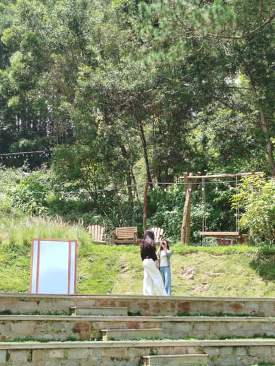 The image depicts an outdoor scene in a lush, green environment, potentially at Cam Ly - Da Lat, Vietnam. In the foreground, a tiered stone structure with steps leads up to a grassy area. A large, rectangular mirror with a wooden frame stands on the grass, reflecting the sky. Further back, two women are visible on the grassy slope. The woman on the left, wearing a black top and white pants, is facing away from the viewer. The woman on the right, in a grey jacket and blue jeans, is holding a camera and appears to be taking a photo.

The background is dominated by dense foliage and trees, suggesting a natural setting. There are wooden chairs and a picnic table visible, along with a wooden swing set. String lights are hung between trees, indicating a possibly recreational or event space. The overall impression is one of a serene, natural landscape with elements designed for relaxation and enjoyment. The sunlight suggests it is daytime.