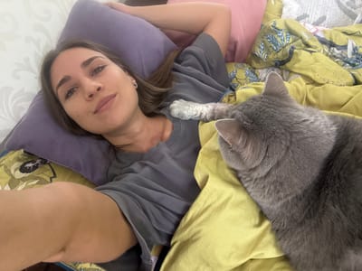 A woman lies on her back on a bed covered in floral-patterned bedding and a yellow blanket. She is using a purple pillow. She is wearing a grey T-shirt. A grey cat is lying to her left, its paw resting on her shirt. The location is Zanevka, Russia.