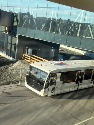 The image shows an airport bus parked next to a glass and steel building. The bus is white with the number "132" visible on its side. A gravel area separates the bus from the building. Part of a wooden structure is visible near the bus. A person in a hardhat can be seen standing next to the bus. The location is not identifiable as the city of Orel, Russia, from visual information provided.