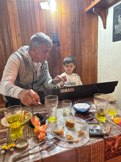 This indoor scene, captured in Mlekarevo, Bulgaria, depicts an older man and a young boy seated at a table laden with food and drinks, alongside an electronic keyboard. The image is oriented sideways, featuring a warm, wood-paneled wall on the left and a lighter-toned ceiling above.
On the left, an older man with short grey hair and a light-colored hooded jacket holds a lit cigarette in his right hand. He is looking towards the young boy. The boy, with dark hair, is to the man's right, wearing a white hoodie with the word "OUR" printed on it. He appears engaged with a black Yamaha electronic keyboard positioned on the table between them, with his hands near the keys. The brand "YAMAHA" is prominently displayed on the instrument.
The table is covered with a patterned brown and white tablecloth. Scattered across it are multiple clear glasses, some containing a yellow liquid. A clear glass plate holds several pieces of partially eaten pastries or sweets. Another plate contains a colorful dish, possibly a salad, alongside black olives. A blue bowl filled with a white granular substance (likely sugar), a pack of cigarettes (faintly showing "MARLBORO" branding), a lighter, a fork, a knife, and a small yellow toy car are also visible on the table. A decorative wooden shelf and a dark rectangular wall hanging with a light-colored relief sculpture are mounted on the wall above the wooden paneling.
The scene suggests a casual and relaxed family gathering, possibly during an evening or late afternoon, given the indoor lighting. The boy seems curious and engaged with the keyboard.