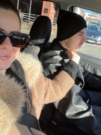 The image depicts two individuals inside a vehicle during daylight in Marquette-lez-Lille, France.
In the immediate foreground, a person with a partially visible face is seen. This individual wears large, dark tortoiseshell sunglasses and a brown coat featuring a light-colored fur trim around the collar. Their left arm, covered by a brown knitted sleeve, extends into the scene.
Seated to the right and slightly behind the first individual, a male is visible in profile. He wears a black knitted beanie and a black leather jacket over a black turtleneck sweater. His right hand is positioned near his neck, and his gaze is directed towards the right, out the vehicle's window.
The vehicle appears stationary. Visible through the windows, in the exterior environment, are a brick building with dark windows and a white railing, a white horizontal barrier pole, and a silver Renault passenger vehicle bearing the French license plate "FG-744-NG". The exterior elements suggest the vehicle is located within a public or commercial area, possibly an entrance or parking lot.