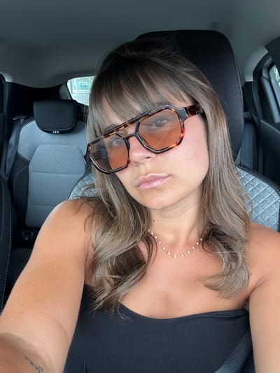 A woman with brown hair and sunglasses is in a car. She is wearing a black top and a pearl necklace. The car has grey seats. No specific location details related to Andrézieux-Bouthéon are discernible.