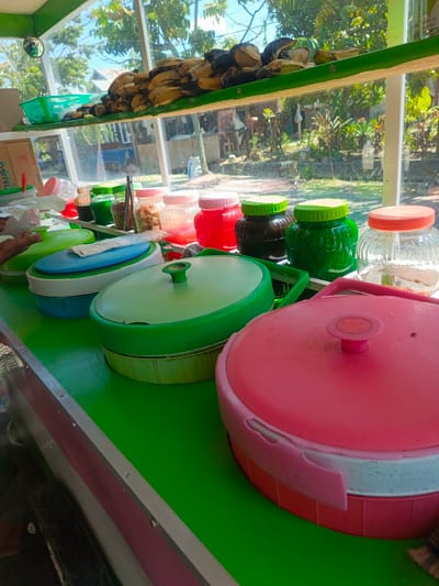 The image shows an outdoor food stall, likely in the city of Luwuk, Indonesia, judging by the presence of ripe bananas, a common fruit in the region. The stall features a vibrant green countertop, upon which several food containers are arranged. Among these are three large, round containers with pink and green lids, suggesting they hold prepared food items. Smaller jars with colorful lids, containing what appear to be liquids or preserved foods, are also visible on a shelf above. In the background, lush green foliage and a bright blue sky indicate a tropical environment. The overall scene depicts a bustling local market setting, with the food stall as the central focus.