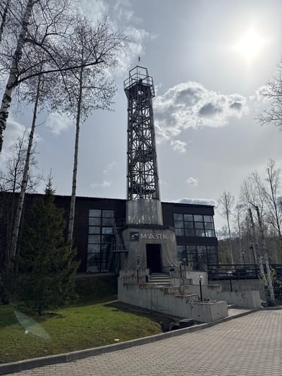 The image depicts a view of the "Mayak" art park in the city of Pavlovka, Russia. A lattice tower stands adjacent to a modern building featuring large windows and concrete elements. Bare trees are visible in the foreground and against a cloudy sky. The surrounding landscape includes a grassy area and a paved walkway.