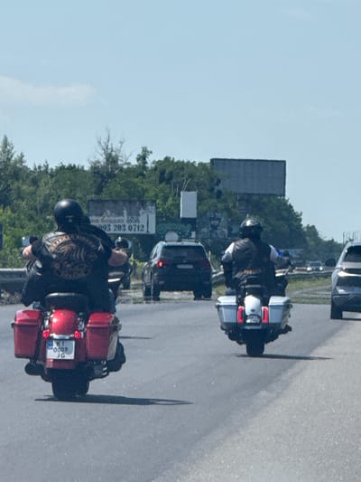 Two individuals riding motorcycles are traveling on a highway. The motorcycle on the left has a red rear section and a license plate reading "KI 0009 JG". The rider is wearing a black helmet and a black shirt with a circular emblem. The motorcycle on the right is silver and white, and its rider is wearing a white shirt and a black vest. In the background, there are trees and a billboard with text and a phone number, possibly indicating an advertisement. Several cars are also visible on the highway, including a dark SUV and a lighter-colored vehicle. The sky is clear and blue with a few wispy clouds. The scene appears to be on a sunny day. The image is a photograph taken from behind the motorcycles.