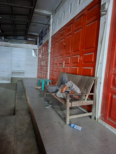 A man sits on a wooden structure next to a red garage door in what appears to be Keudegeudong, Indonesia. He is wearing a grey shirt and pants. A clear plastic water bottle lies on the grey tiled floor next to him. The background includes a white metal fence and black metal racking. A turquoise plastic chair is next to the wooden structure. The scene is rotated for viewing perspective.