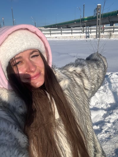 The image displays a young adult female in a close-up, selfie-style photograph taken outdoors in Shul'gino, Russia, on a clear, sunny winter day. The individual is positioned slightly to the left of the frame, smiling faintly with open eyes and visible eyelashes. She wears a pink hood over a white, fluffy hat and a thick, light grey and white faux fur coat. Her long, dark brown hair cascades over her right shoulder and chest, casting a shadow on her left cheek. Her right arm, covered by the fur coat, is extended towards the right side of the frame, suggesting it holds the camera. The background consists of a broad, snow-covered landscape. A small, bare sapling protrudes from the snow in the mid-ground to the right. Further back, an elevated multi-lane highway or bridge, featuring a green barrier along its edge, is supported by concrete pillars. Concrete retaining walls, partially covered in snow, are visible beneath the elevated structure, and streetlights line the highway. The sky is clear blue.