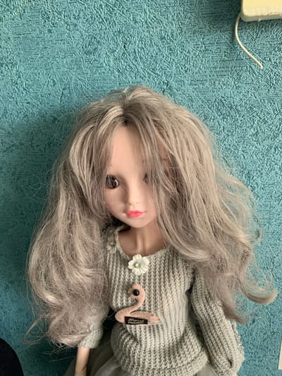 This media file features a doll with long, wavy, light gray hair and brown eyes, lying on a textured blue surface that resembles a carpet or mat. The doll is wearing a gray knitted sweater adorned with a white flower and a pink flamingo-shaped appliqué. A small tag with black text is attached to the sweater. The doll's head is tilted to the side, and its pink lips are slightly parted. To the upper left of the image, a white wire emerges from a white rectangular object. The overall scene is a close-up shot of the doll, with the blue textured surface filling the background. There are no indications of time of day, weather, or a specific event taking place. The location context is Volzhsky, Russia.