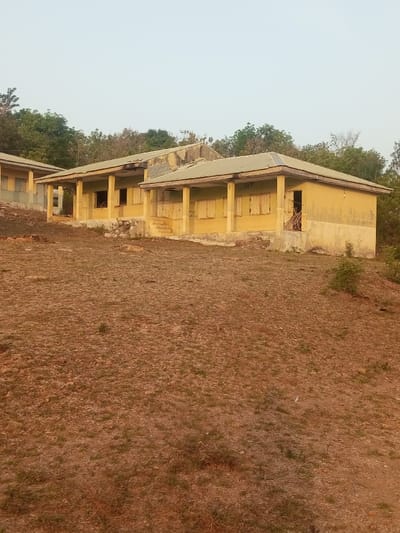 The image displays an exterior view of multiple single-story buildings on a sloping, elevated terrain in Okeni, Nigeria. The foreground consists of dry, reddish-brown earth with sparse patches of green grass and scattered rocks. In the midground, a series of at least three connected, weathered single-story structures with light yellow-ochre painted stucco walls and grey corrugated metal roofs are visible. Each building features a covered porch supported by pillars. Multiple rectangular openings are present; some are boarded with light-colored wooden planks acting as shutters, while others are open. The rightmost building has an open doorway revealing a collection of dry sticks or brush inside. Faint, dark, partially legible text is visible on the exterior wall of the rightmost building, including "S.P.R" and "FOGO LOGO". The buildings are elevated on concrete foundations, with some sections including low steps. The background features a dense line of green trees and vegetation, and the sky above is a uniform pale blue-white. No people or animals are present, and no actions or interactions are depicted.